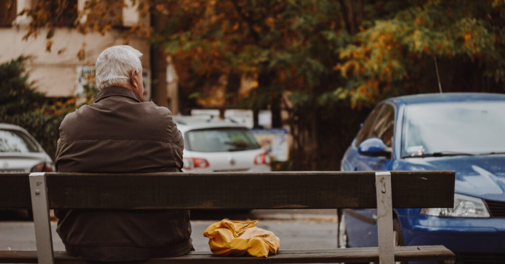 older man sitting on bench with back to camera facing street where cars are parked.